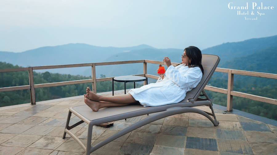 Women in bathrobe relaxing on sun lounger at Grand Palace, Yercaud, on outdoor deck with mountain views, railing, and clear sky in the background.