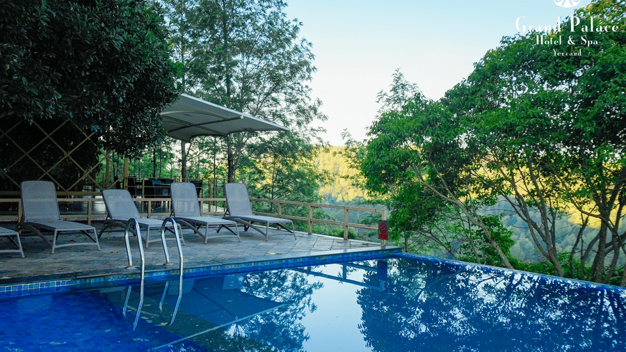Poolside seating at Grand Palace, Yercaud with lounge chairs, table, umbrella, surrounding trees, greenery, pool water, and garden.