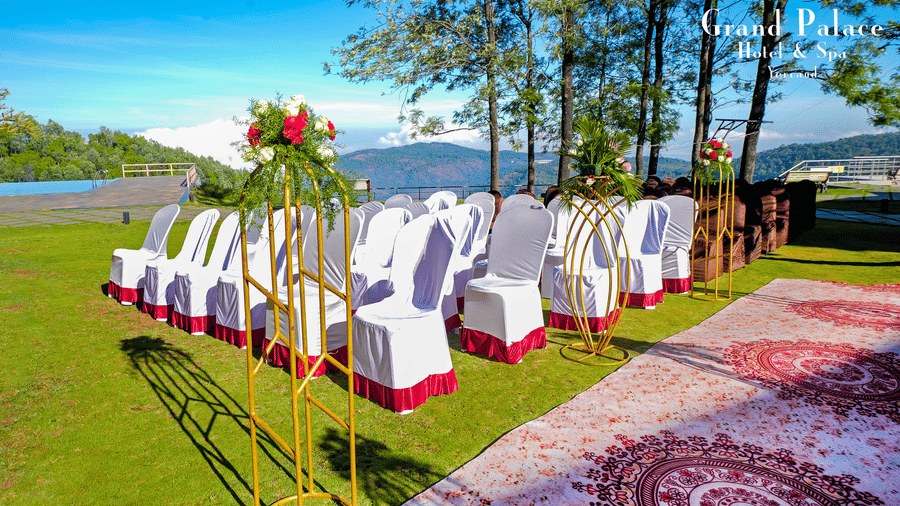 Open-air wedding seating at Grand Palace, Yercaud, showing decorated white chairs, petals, green lawn, aisle runner, trees, floral stands, and sunlight.