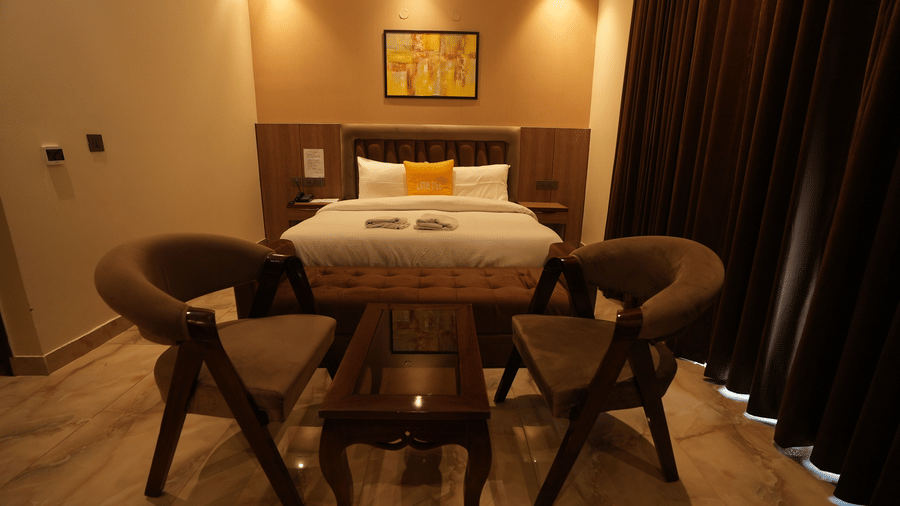 Warmly lit hotel room interior featuring two wooden armchairs and a coffee table in front of the bed.