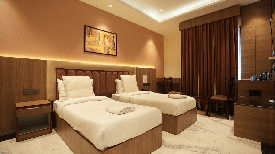 Spacious twin guest room featuring two neatly prepared single beds, warm lighting, and full-length curtains.