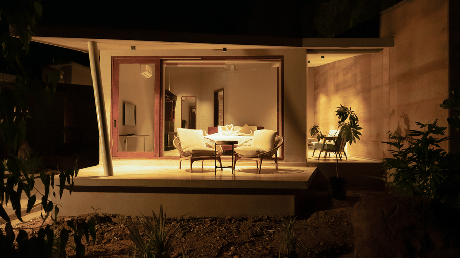 A warmly lit interior space of Gaia Studio with minimalist furniture on the patio and a large opening looking out to a dark exterior at RD's Nature Retreat, Bangalore