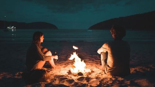 image of two people sitting besides a camp fire on a beach on a late evening
