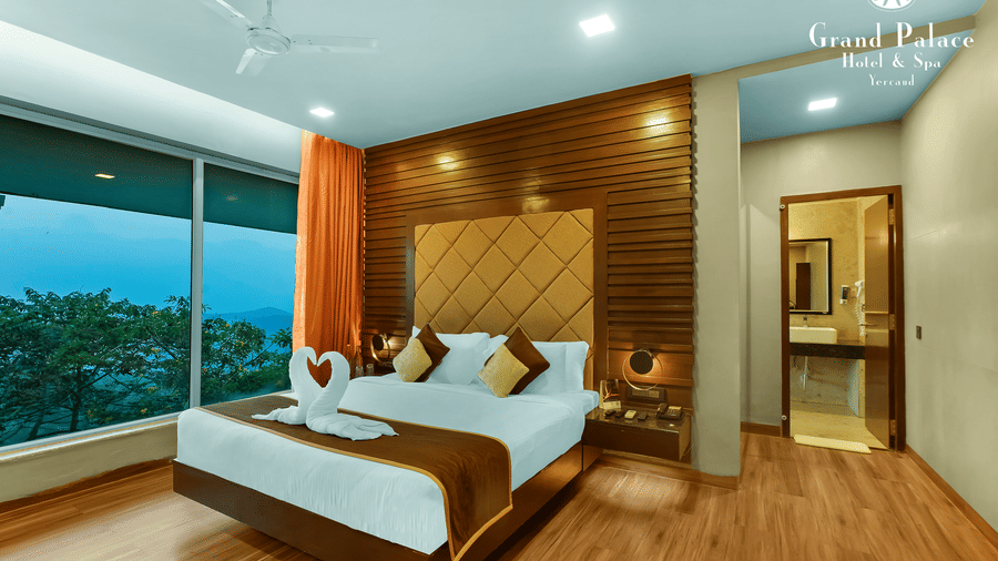 A corner angle Artist Suite at Grand Palace, Yercaud with twin beds, pillows, wooden headboard, curtains, floor lamp, large window, bedside table, and outside greenery visible