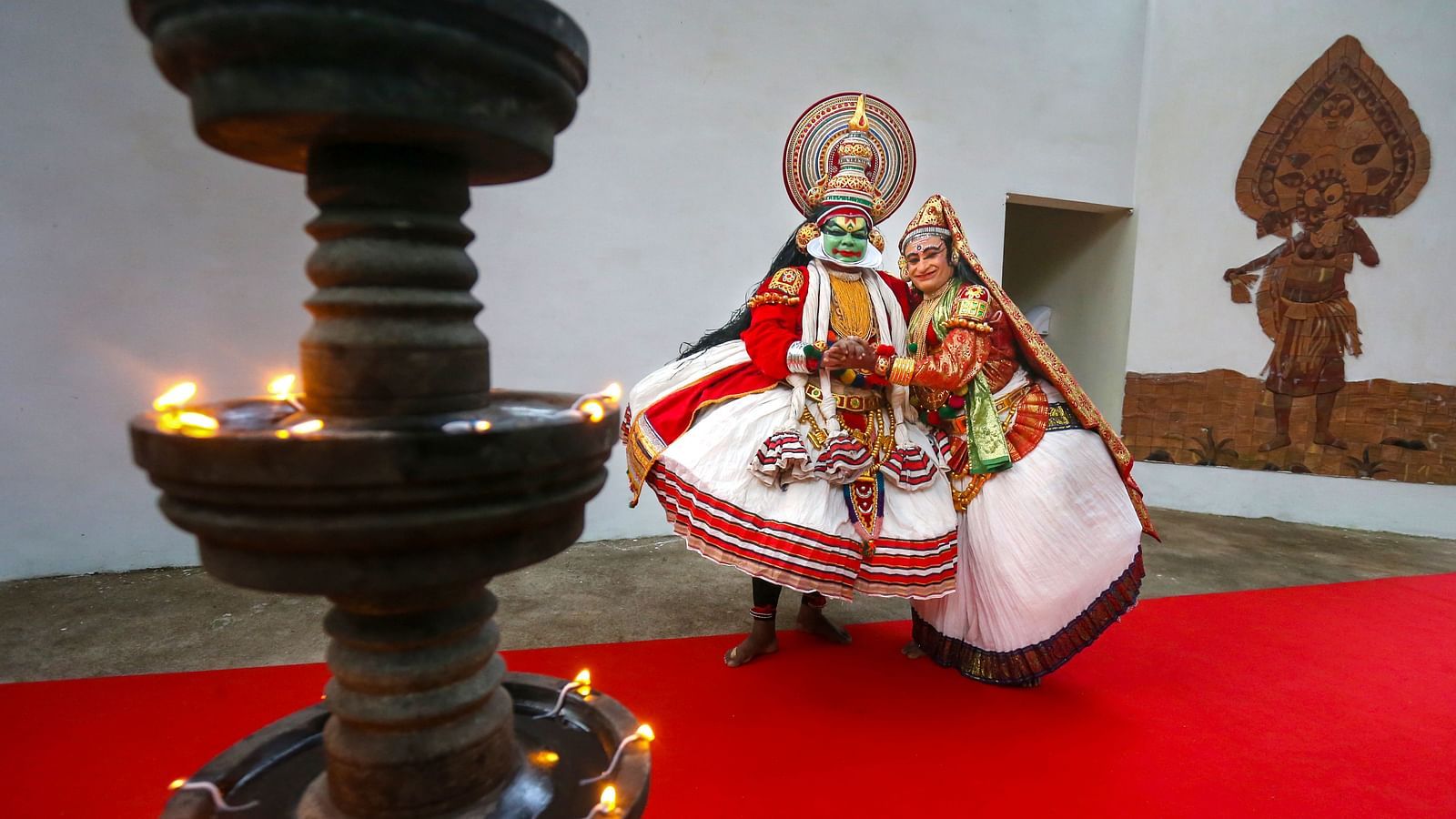 Two people dressed in Kathakali costumes, striking poses near a lamp