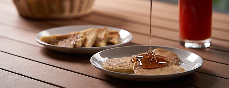 Honey dripping over a plate of pancakes on a wooden table with watermelon juice by side, and a bowl of fruits at The Hamuse Hotel, Kodaikanal.