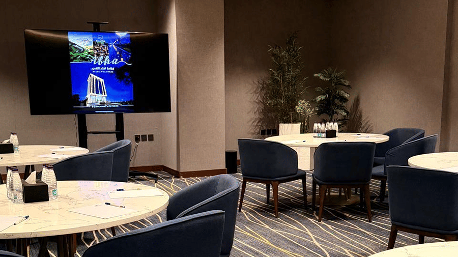 A meeting space at Grand Iva Hotel, Abha featuring round dining tables, cushioned chairs, a display monitor, and floor plants.