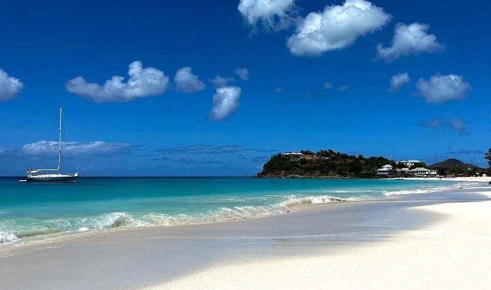 A sailboat drifting on clear turquoise water next to a wide, pristine white sand beach under a bright blue sky with fluffy clouds.