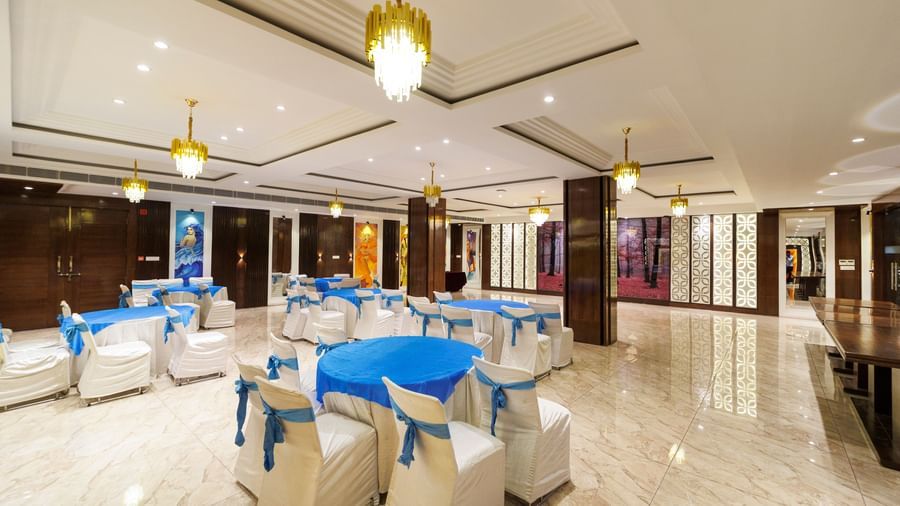 Banquet hall at Inde Reva Resorts Rishhikesh set up with round tables, white chairs, blue covers, and ceiling chandeliers.