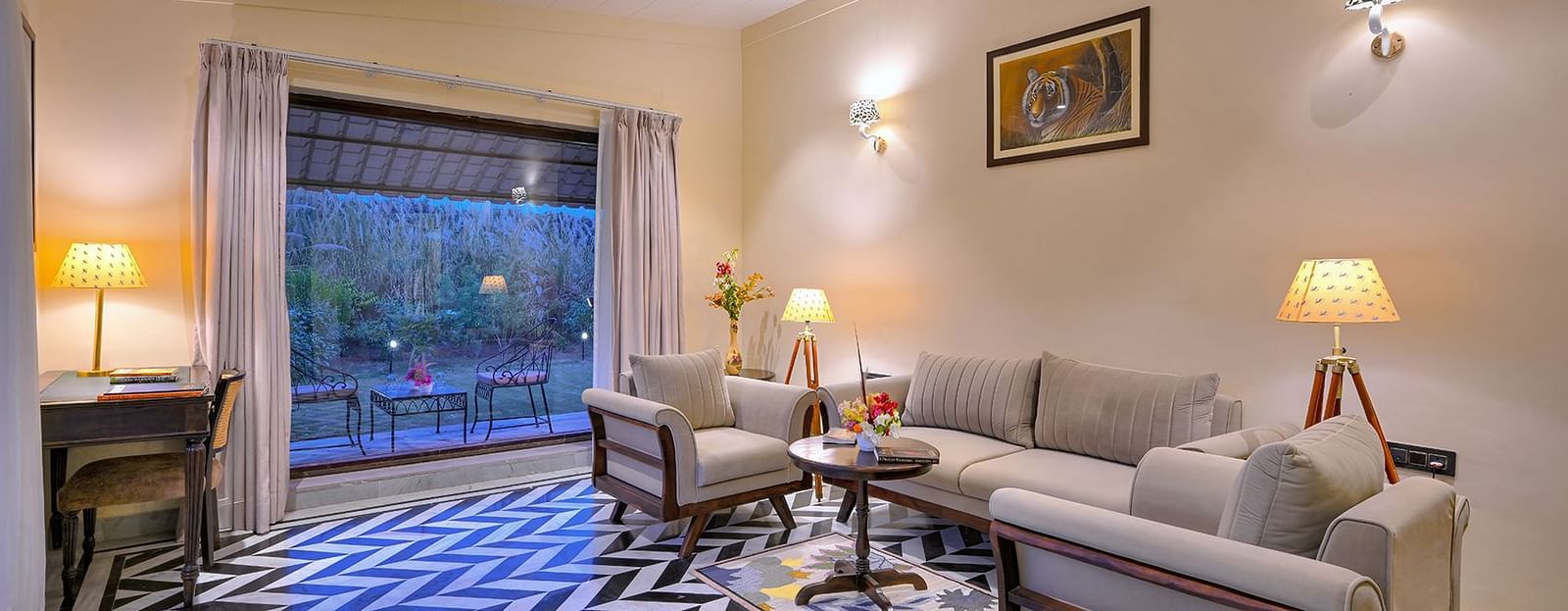 A cosy living room of Superior Cottage with beige sofas, a wooden table, patterned flooring, and a large window overlooking an outdoor seating area - Singhvilas, Ranthambore