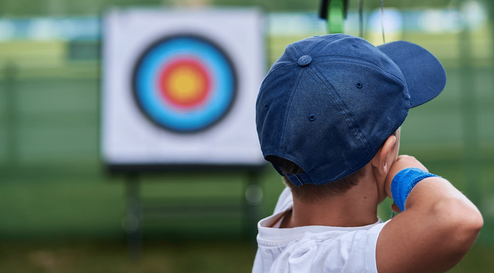 A child wearing a blue cap aims a bow and arrow at a target during archery practice at Nirantara Resort & Spa.