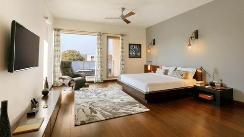 A spacious bedroom with dark wood floors, a low-profile bed, a large window leading to a balcony, a ceiling fan, and a wall-mounted TV - Karma Lakelands