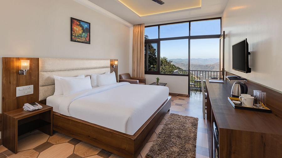 Jungle View Deluxe Room at Pride Ashiyana Resort Mussoorie featuring a plush bed facing the TV mounted on the wall and coffee maker with cups on the table below it. In the background a large window touching the floor and the roof is boasting a majestic vie