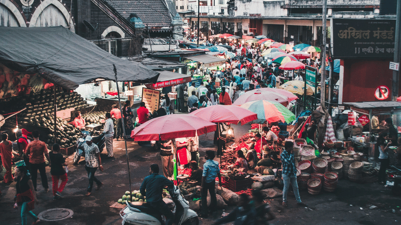 A crowded street market with people holding umbrellas, stalls on both sides, and buildings in the background during rain.