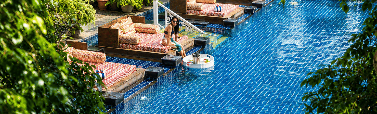 Outdoor pool with blue mosaic tiles, sunken daybeds with patterned cushions, a floating tray and potted palms at MAYFAIR Palm Beach Resort, Gopalpur-on-Sea.