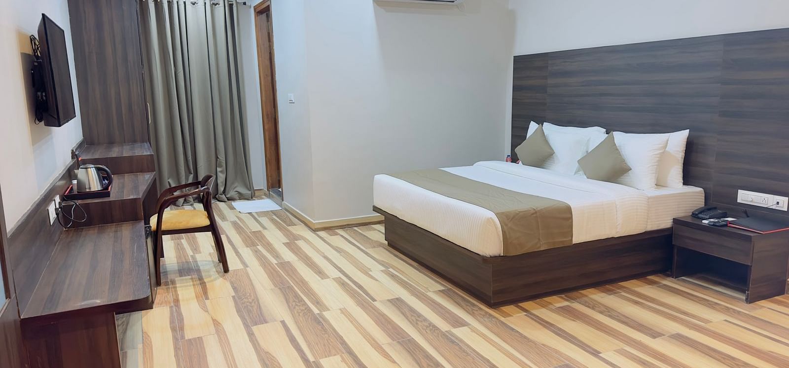A room at Wild Wings Premium features a double bed with beige and white bedding, dark furniture, and wooden flooring - Kankidham Lords Inn