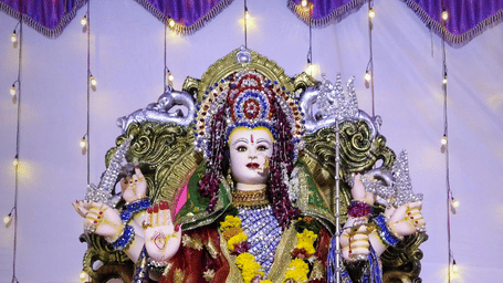 Goddess idol with tiger seat, dressed in red and silver with marigold garlands, surrounded by yellow flowers, candles and fairy lights under a canopy.