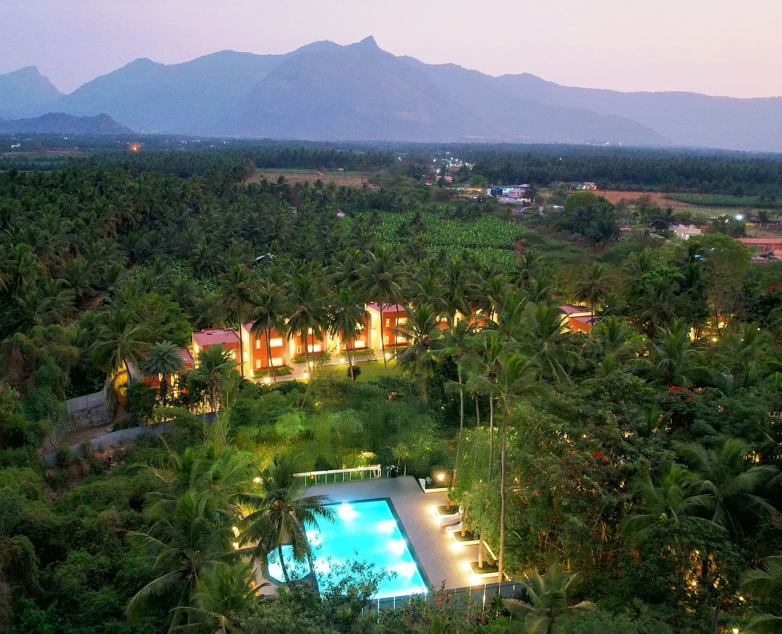 Aerial view of a Mango Hill Resort, Coimbatore with a swimming pool nestled amidst lush greenery and mountains at sunset.