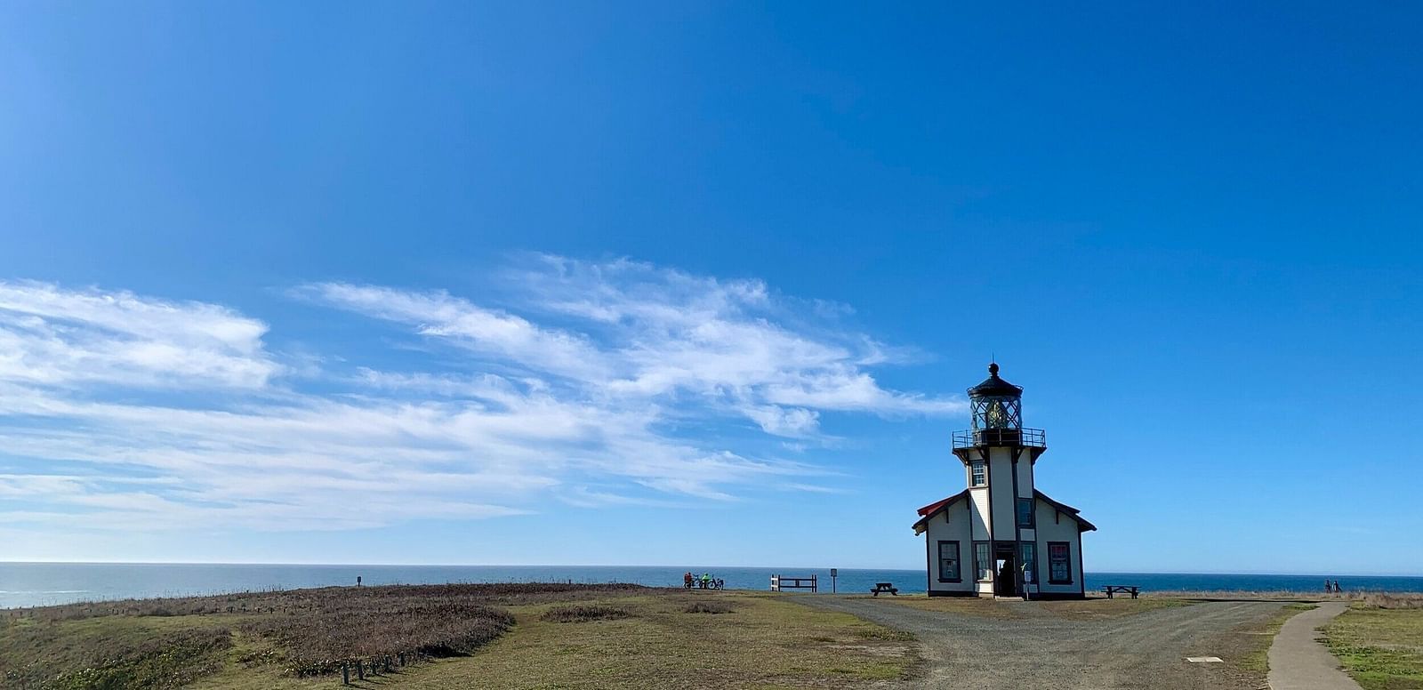 A gravel path leads to a lighthouse perched on a grassy hill overlooking the ocean under a vast sky.