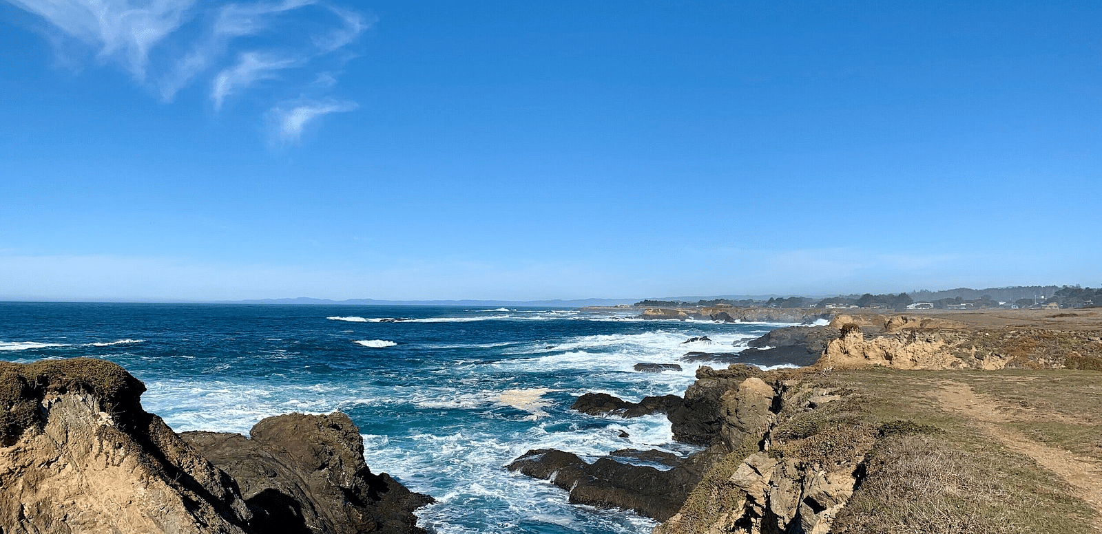 A rocky cliffside overlooks the ocean under a blue sky with wispy clouds, with a path visible along the edge.