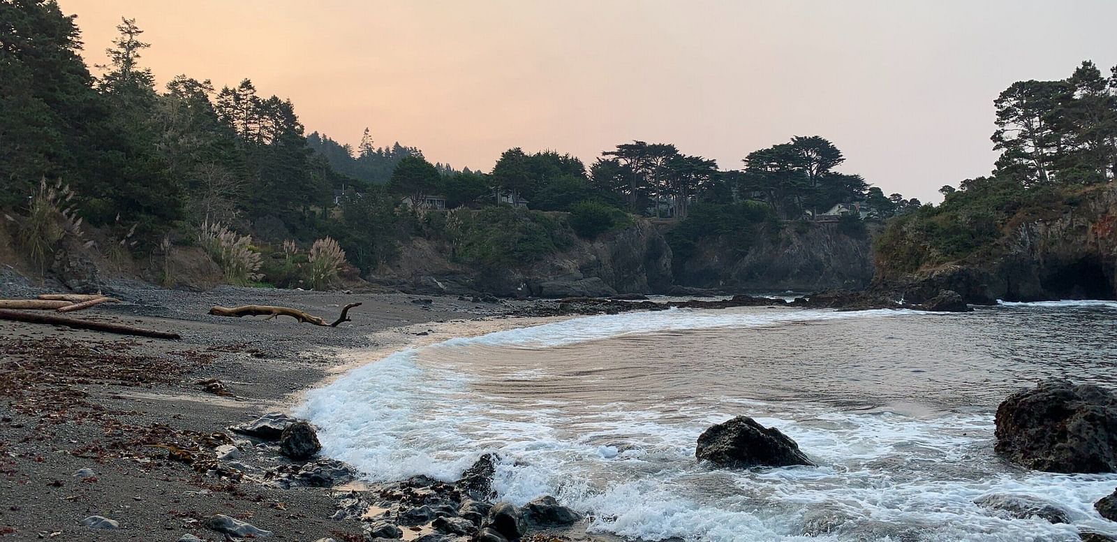 A secluded cove with a dark sandy beach is surrounded by rocky cliffs and lush green trees under a hazy sky.