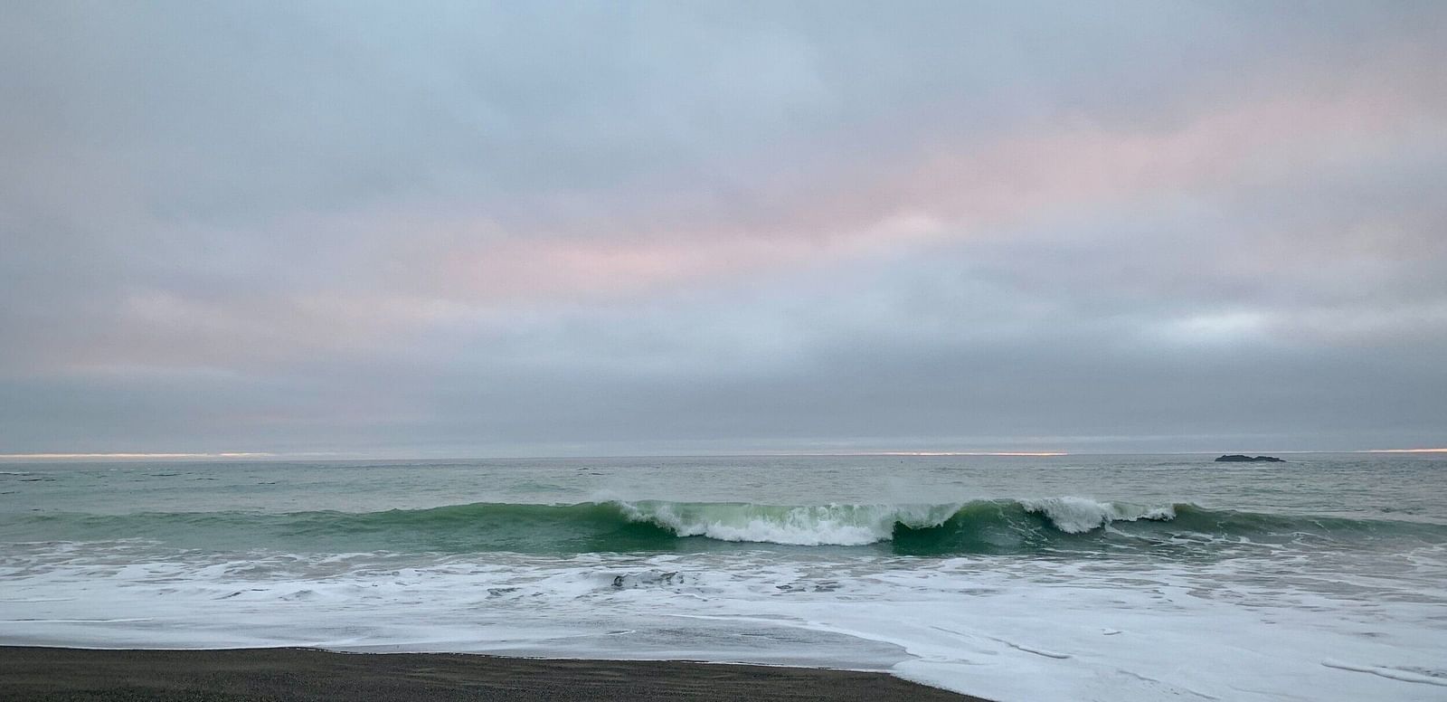 A wave curls and breaks onto a dark sandy beach under a cloudy sky with a faint hint of light.