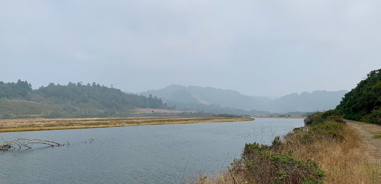 A wide river winds through a marshy landscape with hazy hills in the distance under a cloudy sky - Brewery Gulch Inn & Spa.