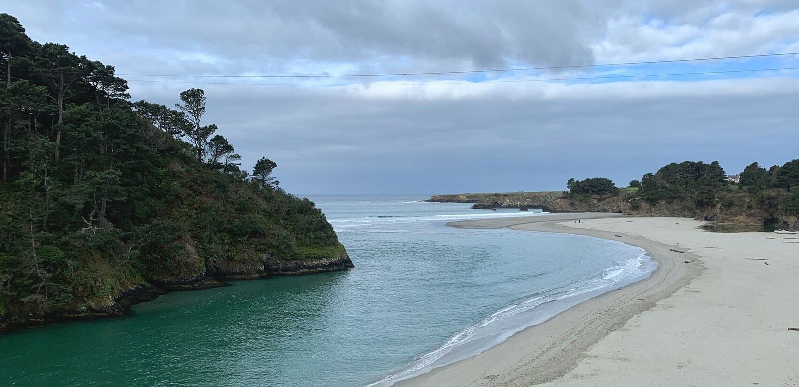 An image of a calm beach with a sandy shore and clear blue-green water, surrounded by cliffs under a cloudy sky
