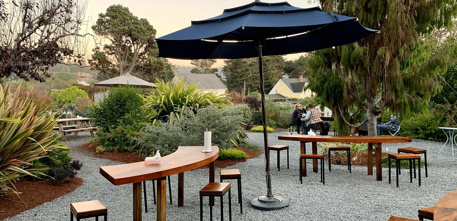 Outdoor patio area with wooden tables, stools, a large umbrella, and various plants under an evening sky.