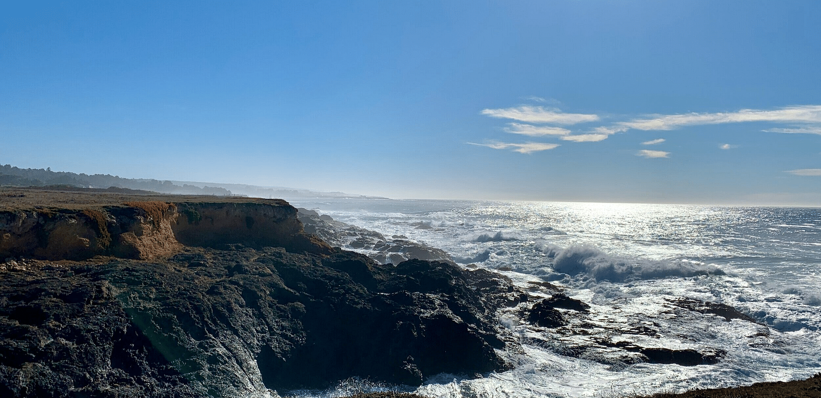 Waves crash against a rocky cliff coastline under a sunny sky, with a sliver of land visible in the distance.