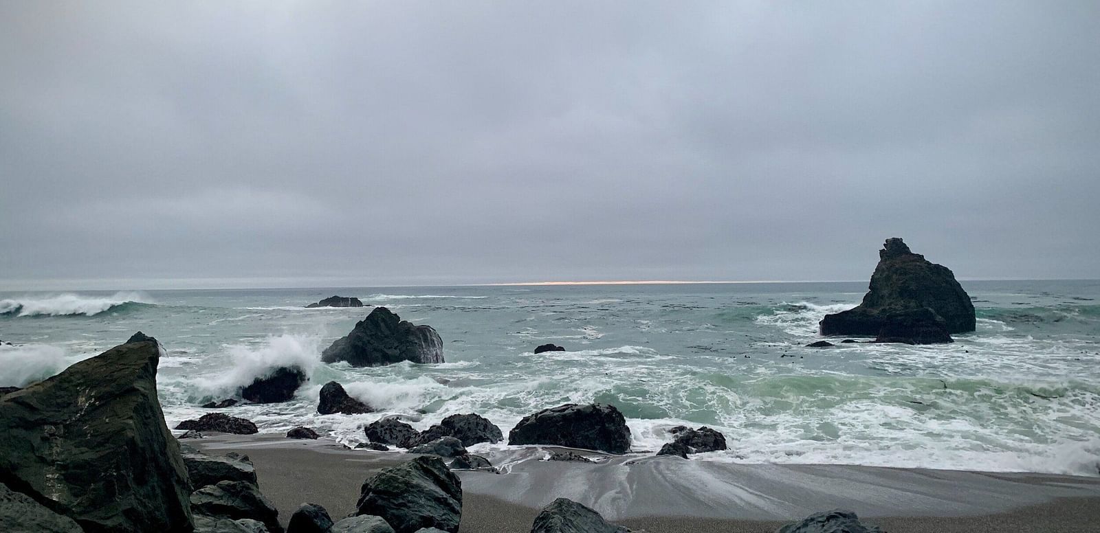 Waves crash over rocks on a pebbled beach under an overcast sky, with a faint light breaking through the clouds on the horizon.