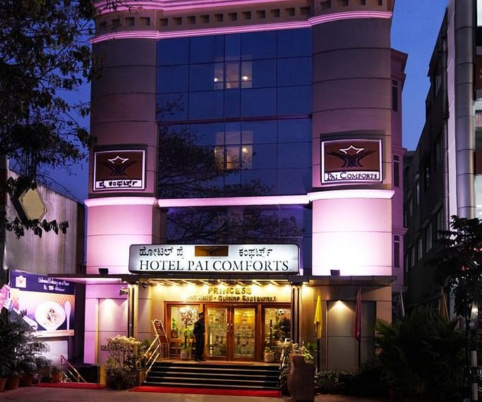 An image of a well lit facade during the night with a backdrop of trees around - Hotel Pai Comforts JP Nagar Bangalore