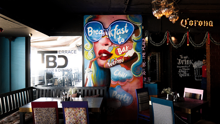 A colourful indoor dining area with a large pop art mural of a woman wearing sunglasses and drinking through a straw. The mural includes the words Breakfast to Bar. The restaurant is called TBD Bar & Dining. 