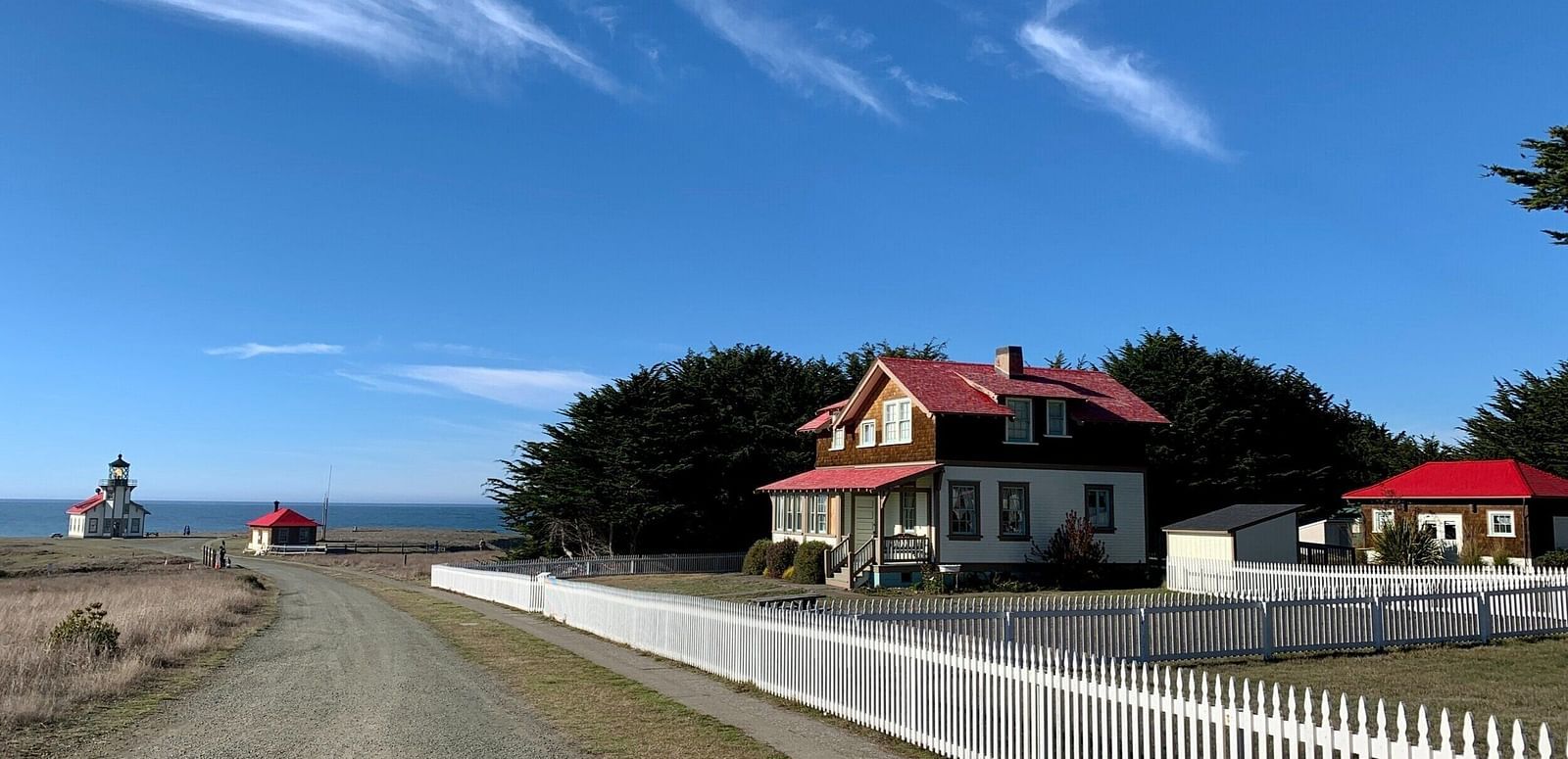 A gravel road leads to a house with a fence, with other buildings and a lighthouse visible in the distance.