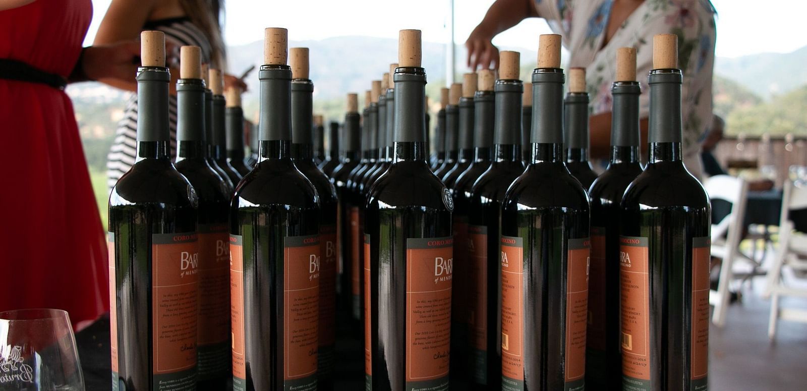 A collection of wine bottles with cork stoppers are arranged closely together, with parts of people visible in the background.