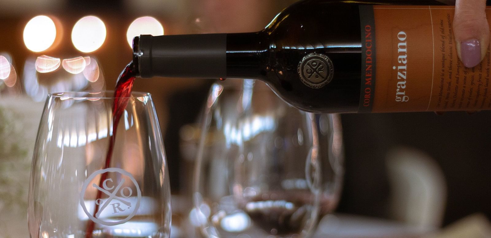 A bottle pours liquid into a glass, with another glass and blurred background elements visible.