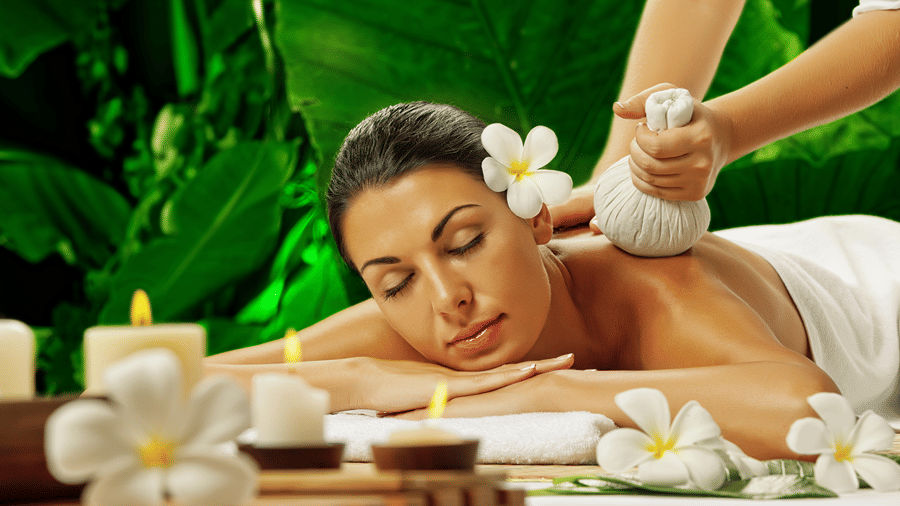 Woman receiving a herbal compress spa massage therapy session surrounded by lit candles and frangipani flowers.