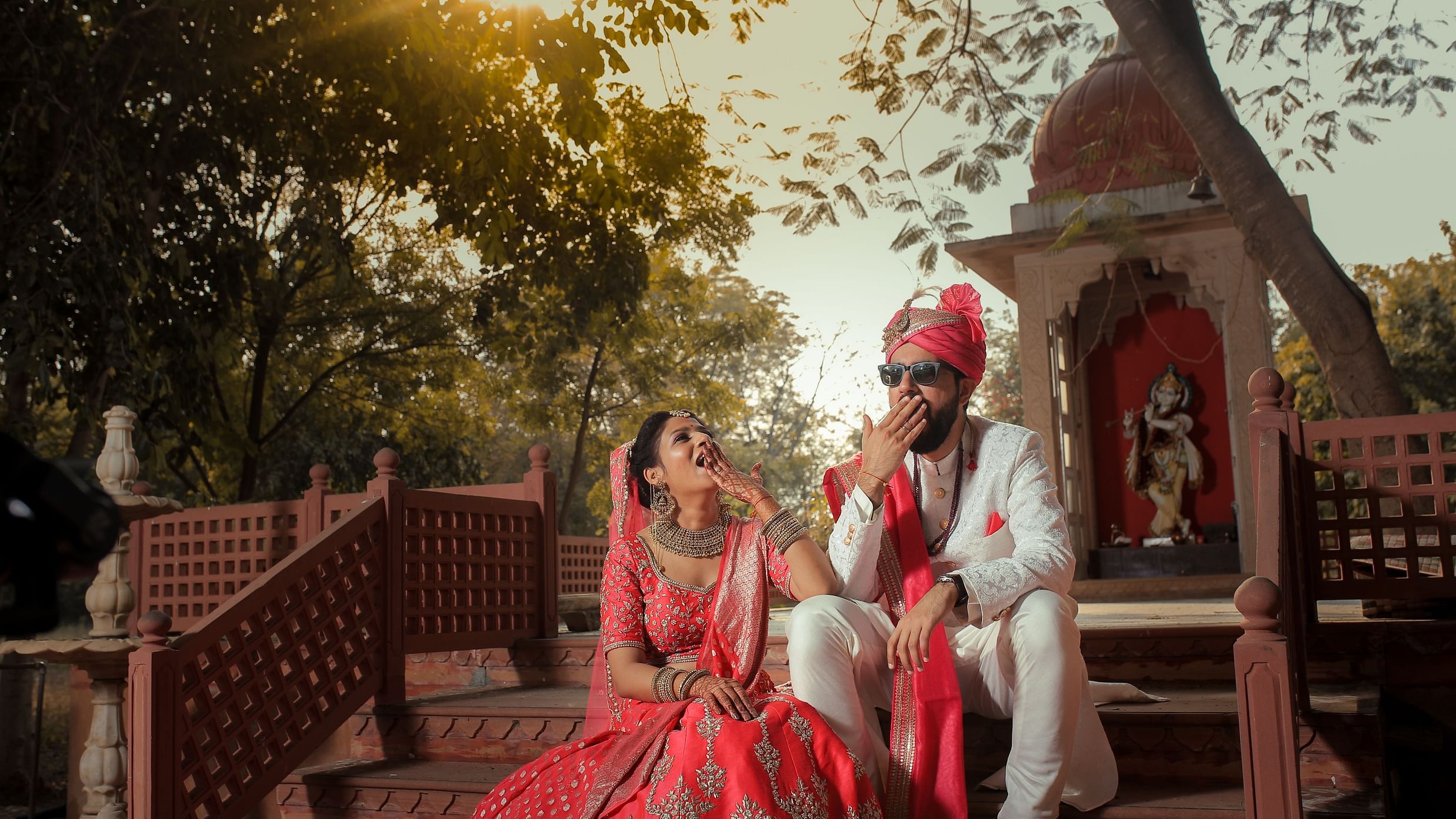 A newlywed couple in wedding attire sits on temple stairs, laughing and playfully covering their mouth at a Jaipur's Top Wedding Resort - Umaid Palace - A Lakeside Heritage Palace Resort
