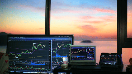 A close up of three monitors with stock trading graph on each of them and different hues on the sky in the background.