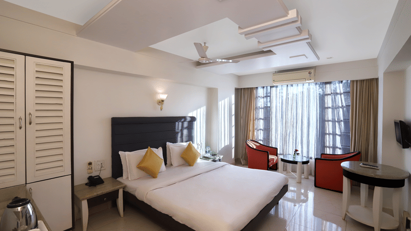 Spacious, well-lit hotel room at Hotel Kohinoor Highway, Dapoli with a large double bed, bright cushions, modern furnishings, balcony seating, and natural light streaming through curtains