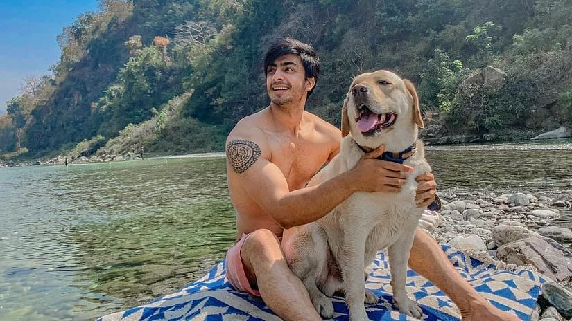 A man sits on a blanket by a river with a dog beside him