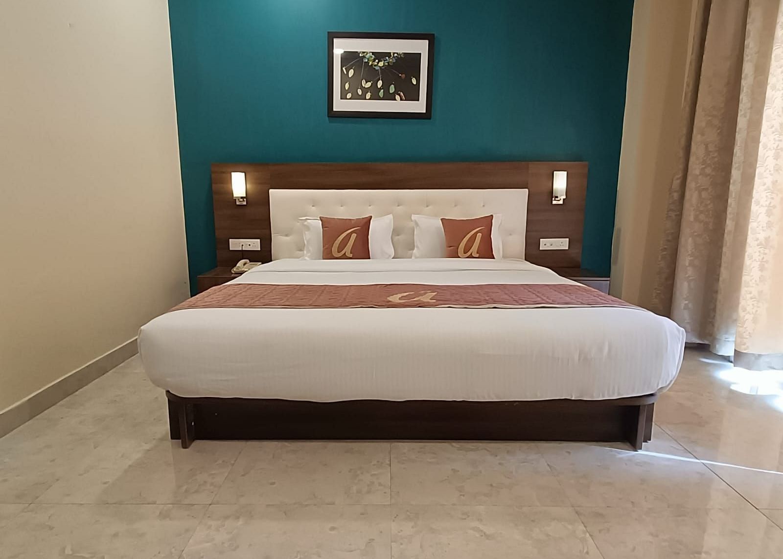   A neatly made hotel bed with a white duvet and maroon accents, pillows against a dark teal wall with a framed picture, and beige curtains.