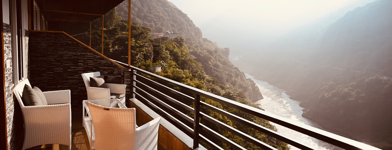 Shot of the mountains and river flowing from the balcony of the hotel featuring two white chairs and table in the morning | The Tattva Devprayag