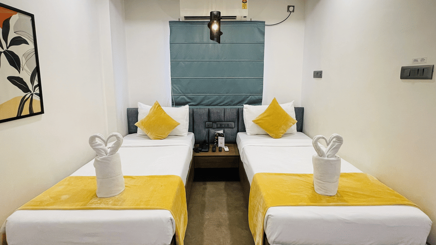 Close view of twin beds at Hotel Kyzen Smart Stays Madhapur with yellow cushions, swan towel folding, teal headboard wall panel, and bedside lighting.