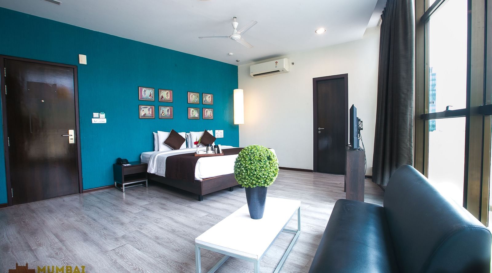 Spacious hotel room with a seating area, wooden floor, turquoise accent wall with art, a white coffee table, and a window.