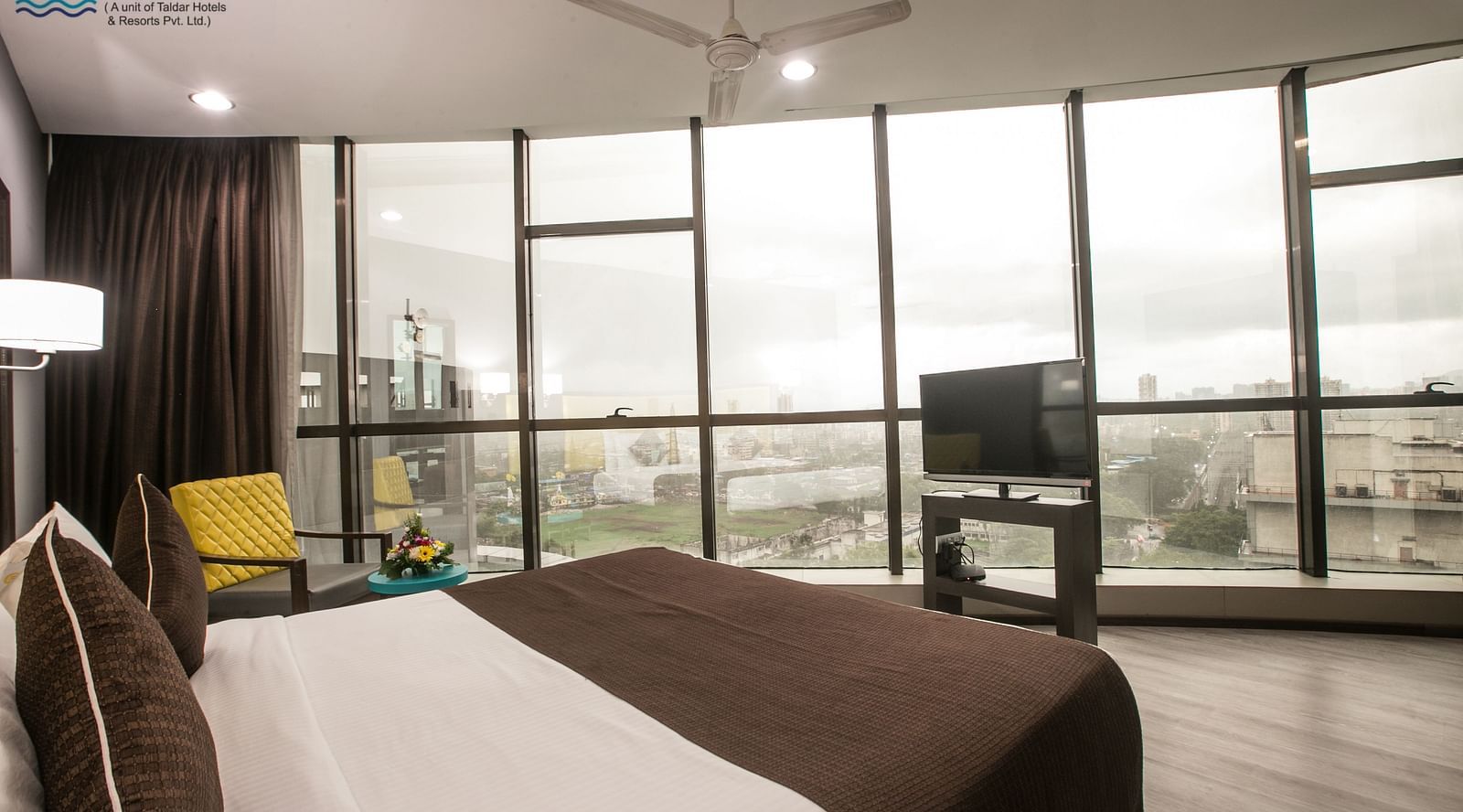 Hotel room with a large window offering a city view, a bed with brown bedding and decorative pillows, and a flat-screen TV.
