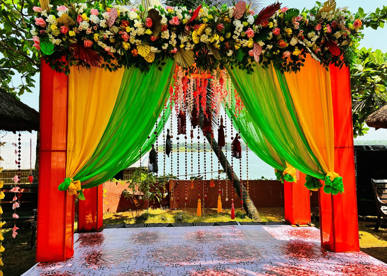 Wedding mandap decorated with orange and green drapes, flowers, and hanging garlands at Paradise Lagoon Resort, Udupi.