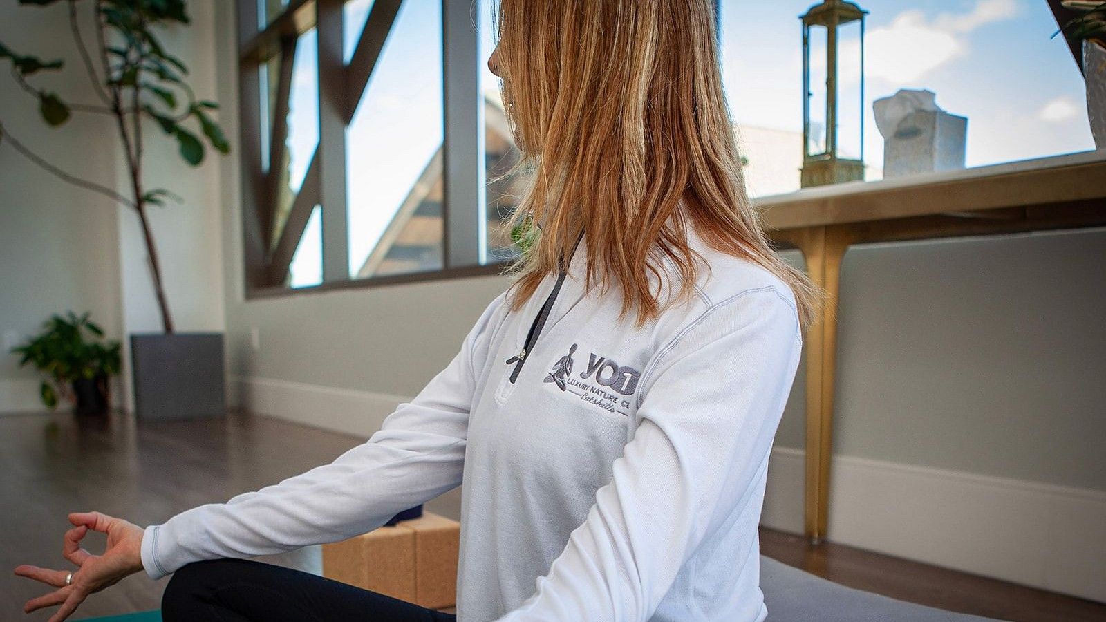Woman seated cross-legged on a yoga mat indoors, wearing a white top, practicing meditation with hand in Gyan mudra near large windows at YO1 Longevity and Health Resorts, Catskills.