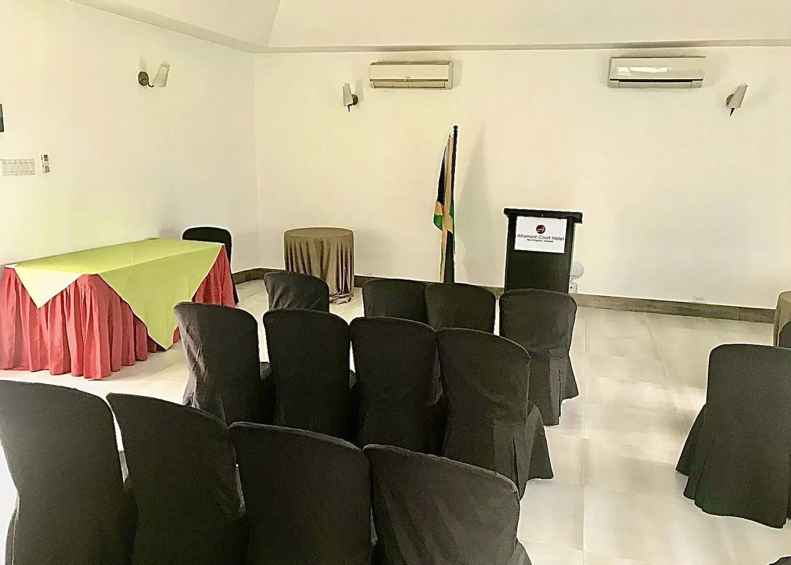 An empty conference room features rows of black chairs facing a table with a television screen and a Jamaican flag | Altamont Court, Jamaica