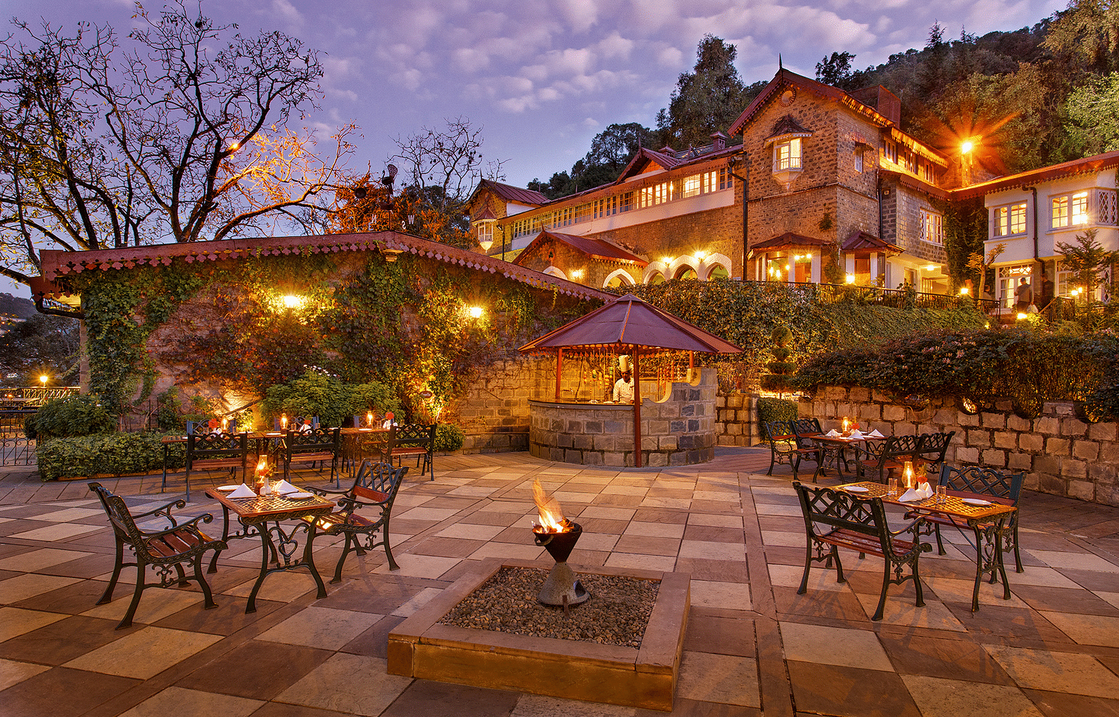 A beautiful outdoor dining area at the naini retreat, nainital. There are tables and chairs set up on a stone patio, with a fireplace in the center. The building is lit up at dusk, creating a warm atmosphere. 